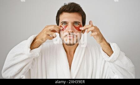 Hübscher bärtiger junger Mann in einem weißen Gewand, der rote Flecken unter den Augen vor einem isolierten weißen Hintergrund aufträgt. Stockfoto