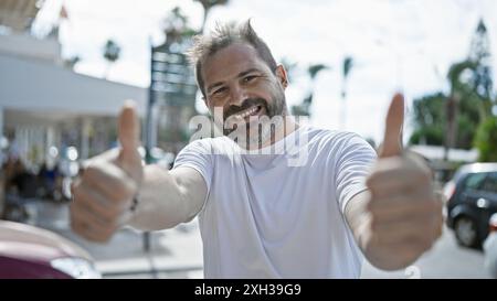 Ein gutaussehender reifer hispanischer Mann mit grauen Haaren, der die Daumen nach oben auf einer sonnigen Straße im Freien hält. Stockfoto