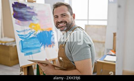 Ein lächelnder bärtiger Mann mit grauen Haaren auf Leinwand in einem hellen Atelier, der seine schöne, reife Künstlerpersönlichkeit zeigt. Stockfoto