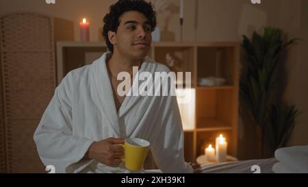 Hübscher junger hispanischer Mann in einem weißen Gewand mit einem gelben Becher in einem ruhigen Spa-Raum mit brennenden Kerzen, der ein ruhiges und Wellness-fokussiertes atmos schafft Stockfoto