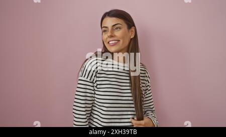 Eine lächelnde junge hispanische Frau in einem gestreiften Hemd posiert vor einem schlichten rosa Hintergrund für ein lebendiges Porträt. Stockfoto