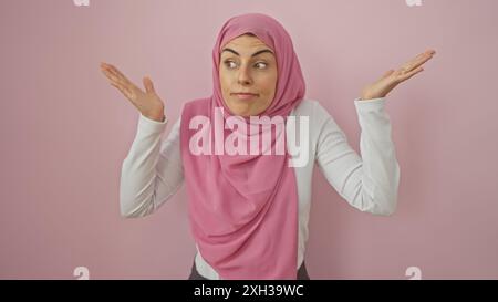 Eine verwirrte Frau in einem rosafarbenen Hijab macht eine fragende Geste vor einem schlichten rosa Hintergrund. Stockfoto
