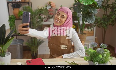 Lächelnde Frau mit Hijab macht Selfie in einem mit Pflanzen gefüllten Innenraum und zeigt einen Lifestyle-Moment zu Hause. Stockfoto
