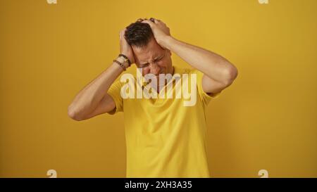 Ein hispanischer Mann mittleren Alters, der ein gelbes Poloshirt trägt, hält seinen Kopf frustriert vor einem leuchtend gelben Hintergrund. Stockfoto