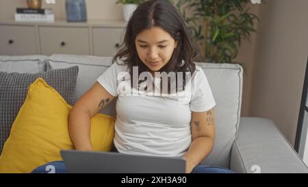 Eine schöne junge hispanische Frau arbeitet an ihrem Laptop im gemütlichen Wohnzimmer ihrer Wohnung. Stockfoto