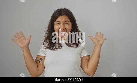 Eine junge, brünette hispanische Frau lächelt begeistert und hebt ihre Hände aufgeregt vor einer weißen Hintergrundwand. Stockfoto