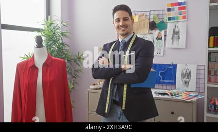 Hübscher Mann mit Bart in Schneiderei Arme gekreuzt, lächelnd in Atelier mit Schaufensterpuppe und Stoffmustern. Stockfoto