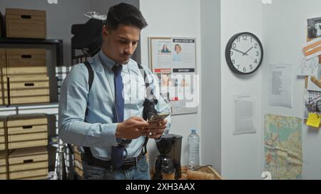 Gutaussehender hispanischer Mann im Detektivbüro, der Nachrichten am Telefon liest, umgeben von Tafel und Karte. Stockfoto