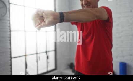 Kahler, hispanischer Mann mittleren Alters dehnt sich in einem hell beleuchteten Fitnessstudio, trägt ein rotes Hemd und einen Fitness-Tracker am Handgelenk. Stockfoto