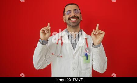 Junger hispanischer Mann mit Schnurrbart und Brille in weißem Labormantel, Lächeln und Gesten mit beiden Händen vor rotem Hintergrund Stockfoto