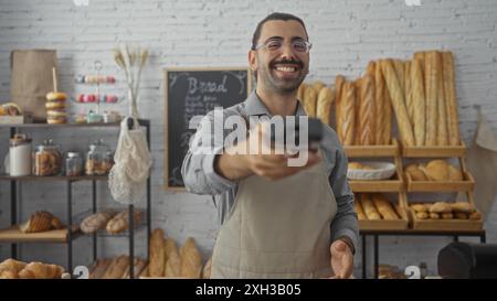 Junger Mann in einer Bäckerei lächelt und hält ein Zahlungsterminal, umgeben von Brot und Gebäck drinnen Stockfoto