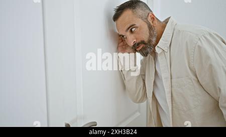 Gutaussehender hispanischer älterer Mann mit grauem Bart blickt neugierig durch die Eingangstür in einladender Mode. Stockfoto