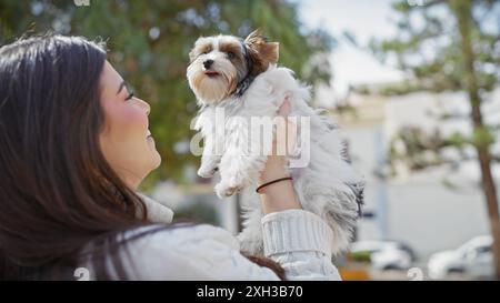 Eine lächelnde junge hispanische Frau genießt einen sonnigen Tag draußen und hält ihren entzückenden biewer yorkshire Terrier auf einer städtischen Straße. Stockfoto