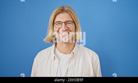 Ein lächelnder Mann mit langen blonden Haaren und Brillen, der sich vor blauem Hintergrund posiert. Stockfoto