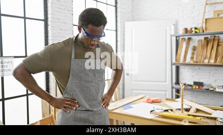 Ein junger afroamerikaner in einer Werkstatt, der eine Schutzbrille und eine Schürze trägt, konzentriert sich intensiv auf ein Tischlerprojekt. Stockfoto