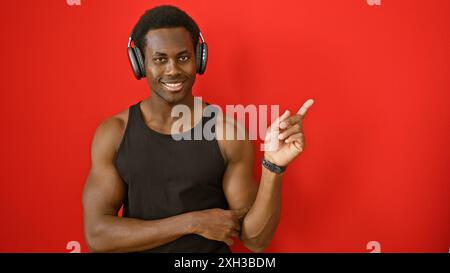 Ein lächelnder junger afroamerikaner, der auf einen roten Hintergrund zeigt und Kopfhörer trägt. Stockfoto