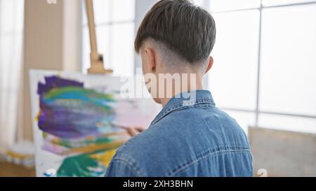 Ein junger Mann in Denim schaut auf ein farbenfrohes abstraktes Gemälde in einem hellen Atelier-Setting. Stockfoto