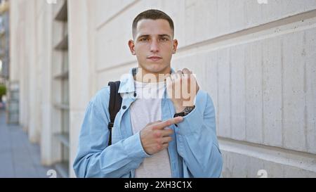 Hispanischer junger Mann in lässiger Kleidung, der auf seine Uhr auf einer Stadtstraße zeigt, pünktlich und bereit aussieht. Stockfoto