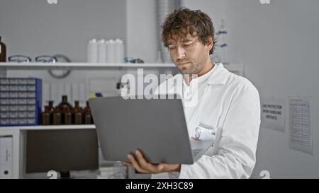 Hispanischer Mann mit Bart, der im Labor an einem Laptop arbeitet und drinnen weißen Labormantel trägt. Stockfoto