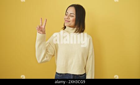 Eine junge hispanische Frau in einem Pullover lächelt und gibt Frieden vor gelbem Hintergrund. Stockfoto