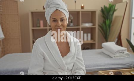 Frau mittleren Alters, die ein entspannendes Spa-Erlebnis in einem ruhigen Schönheitssalon genießt, gekleidet in einem weißen Mantel mit einem Handtuch um den Kopf, umgeben Stockfoto