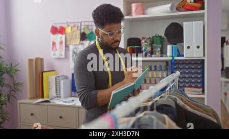 afroamerikanischer Schneider macht Notizen in einem farbenfrohen Atelier, umgeben von Stoffen und Designskizzen. Stockfoto