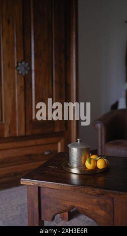 Eine alte Kaffeekanne und frische Orangen auf einem Messingtablett, auf einem Holztisch in einem rustikalen Interieur mit Ledermöbeln. Stockfoto