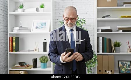 Ein reifer bärtiger Mann im Anzug überprüft sein Smartphone in einem modernen Büro. Stockfoto
