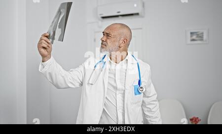 Reifer bärtiger Mann in einem weißen Mantel, der ein Röntgenbild in einem hell beleuchteten Krankenhauszimmer untersucht. Stockfoto