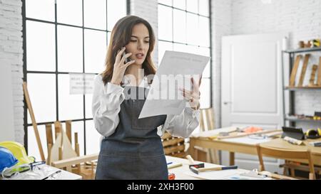 Eine hispanische Architektin, die im hellen Büro beim Telefonieren Pläne überprüft Stockfoto