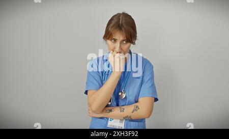 Nachdenkliche Krankenschwester mit Tätowierungen isoliert gegen eine weiße Wand, die professionelles Gesundheitsverhalten ausstrahlt. Stockfoto