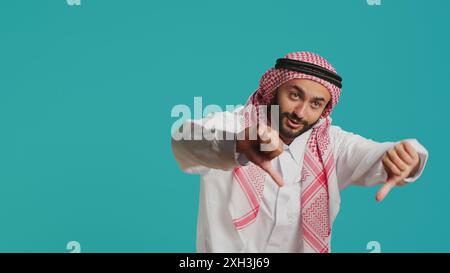 Eine arabische Person in historischem Kleid und Kufiyah gibt die Daumen nach unten und zeigt Unzufriedenheit mit dem, was sie sieht. Im Studio zeigt islamische Erwachsene eine deutliche Ablehnung und ein negatives Signal. Stockfoto