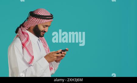 Arabische Person, die mobile Videospiele mit angespanntem Ausdruck auf dem Smartphone spielt, trägt islamisches Kopftuch und Dumbe. Muslimischer Gamer genießt Online-Gaming-Erlebnis über Telefon-App im Studio. Stockfoto