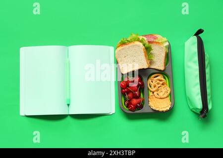 Bleistiftetui, offenes Notizbuch, Lunchbox mit Beeren und Sandwiches auf grünem Hintergrund. Draufsicht Stockfoto