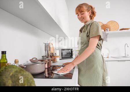 Eine junge Frau, die in der Küche den Elektroherd mit einem Papiertuch abwischt Stockfoto