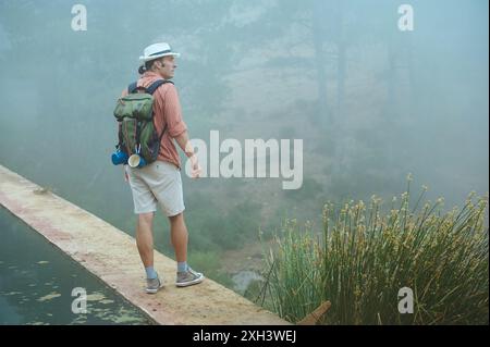 Mann mit Hut und orangefarbenem Hemd, mit Rucksack, stehend auf einem Weg in einer nebeligen Waldumgebung. Konzept: Abenteuer, Natur, Erkundung. Stockfoto