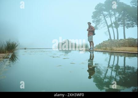 Ein Alleinreisender steht an einem nebeligen Tag in der Nähe eines ruhigen Wasserkörpers. Bäume spiegeln sich im Wasser und schaffen eine ruhige, friedliche Atmosphäre. Das Bild erinnert an einen Sen Stockfoto