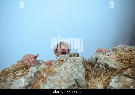 Ein Mann, der extreme Angst hat, während er sich an einer felsigen Klippe festhält. Das Bild fängt intensive Emotionen und das Abenteuer und die Gefahr des Kletterns in na ein Stockfoto