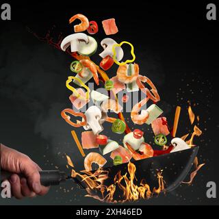 Mann wirft Zutaten in Wok über Feuer auf schwarzem Hintergrund, Nahaufnahme Stockfoto