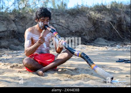 Ein Aborigine-Mann in traditioneller Kleidung und Körperfarbe sitzt am Strand von K'gari und spielt ein Didgeridoo Stockfoto