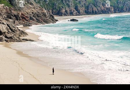 Junge Frau, die allein zu Fuß auf die Meereswellen am weißen Sandstrand der Porthcurno Bay in Cornwall, England, ansieht Stockfoto