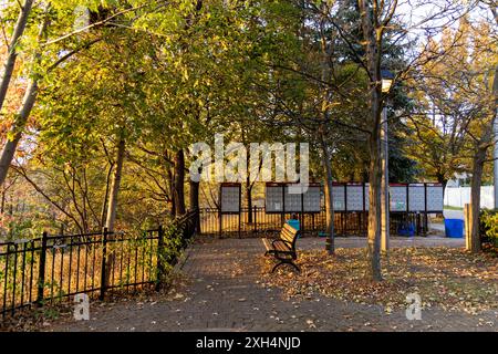 Herbstliche Parkszene - lebhaftes Laub - leere Bank - gepflasterter Fußweg - ruhige städtische Grünfläche - Briefkästen im Hintergrund. Aufgenommen in Toronto, Kanada. Stockfoto