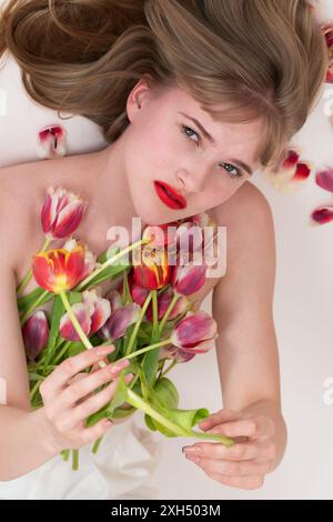 Junge Frau mit Frühlingstulpen, die auf der Brust liegen und sie bedecken, in die Kamera schauen. Blonde Frau, die Blumen hält, während sie sich auf weißem Hintergru legt Stockfoto