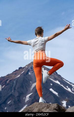 Rückansicht einer Frau, die Yoga praktiziert, auf einem felsigen Berggipfel mit einem Vulkan im Hintergrund. Eine formschöne Frau gleicht den Körper auf einem Bein aus. Yogalehrer Stockfoto