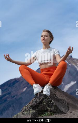 Yogalehrerin sitzt in entspannter Pose auf Stein, nachdem sie einen hohen Berg mit Vulkankegel im Hintergrund bestiegen hat. Blonde Frau mit kurzen Haaren Stockfoto