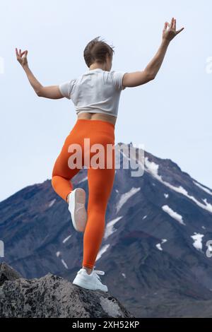 Atemberaubende Rückansicht einer Yogalehrerin, die auf einem felsigen Berg steht und ihren Körper auf einem Bein balanciert, während sie Yoga mit Vulkan macht Stockfoto