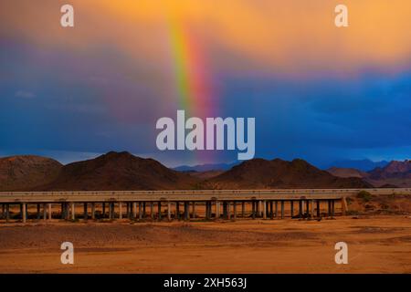 Helle Regenbogenbögen über dramatische Wüstenhügel und einen erhöhten Highway in Kalifornien. Kontrast zwischen lebhaften Farben, rauem Gelände und gemischten Himmelsfarben c Stockfoto