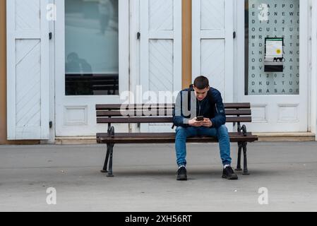 Junger Mann, der allein auf einer Bank sitzt, auf seinem Handy schreibt oder vielleicht im Internet surft. Novi Sad April 2024. Stockfoto