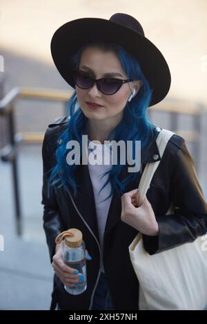 Eine junge Frau mit blauen Haaren, die Hipster-Hut und Lederjacke trägt, spaziert durch die Stadt Stockfoto