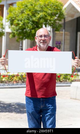 Ein Mann in einem roten Hemd hält ein leeres Schild. Das Schild ist weiß und hat keine Aufschrift. Der Mann lächelt und ist glücklich. Die Szene spielt in einem PA Stockfoto